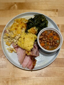 White plate with sliced ham, servings of mac and cheese, collard greens, black eyed peas and cornbread