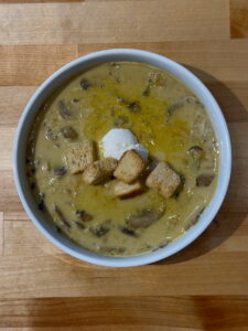 White bowl of creamy wild mushroom and rice soup with a dollop of ricotta cheese and five croutons on top