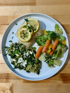 White plate with cooked lemon pepper cod and lemon slices, steamed vegetables and spinach orzo feta pasta