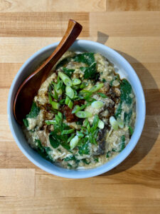 White bowl with the spinach mushroom egg congee finished with a swirl of sesame oil, reduced sodium soy sauce, chili paste and chopped green onions. A wooden spoon is place in the congee at the top of the bowl.