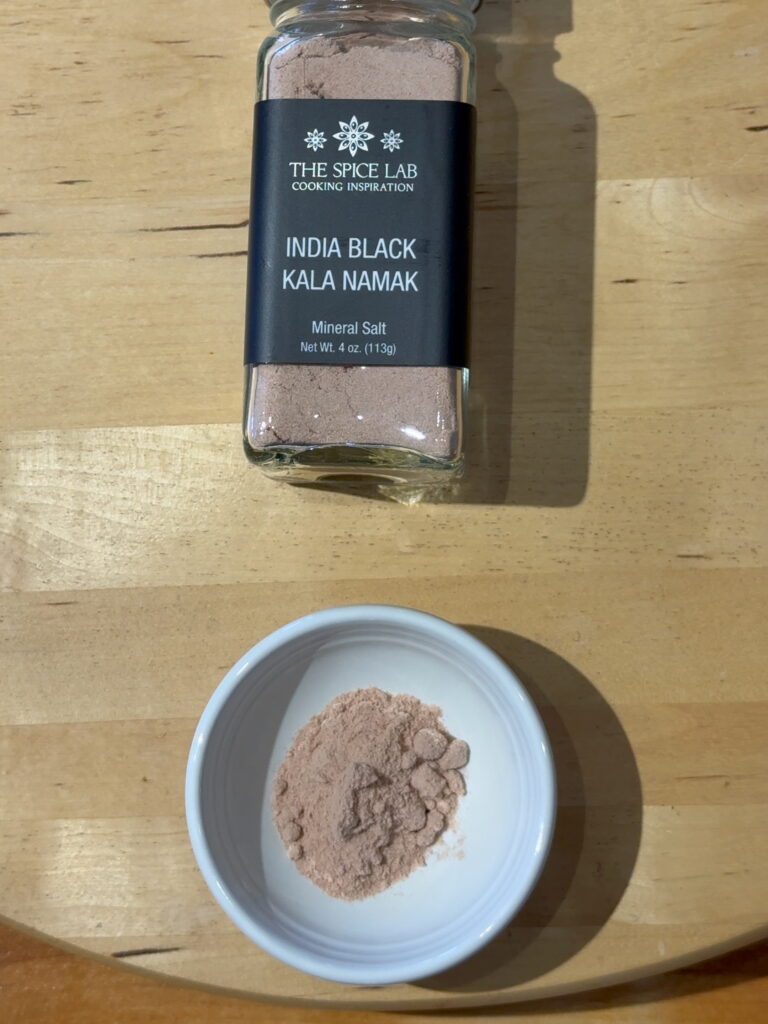 Jar of India Black Kala Namak salt with a small white dish containing the black kala namak salt