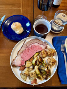 A slice of prime rib steak on a white plate with roasted Brussels Sprouts, stuffed mushrooms and potatoes on a white plate. A sliced roll on a small blue plate. A cup of au jus gravy. A glass of wine and a glass of water.