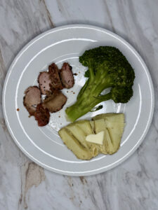 White plate with sliced grilled pork tenderloin, half a baked potato cut open with butter on top and a large spear of steamed broccoli