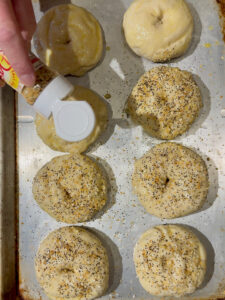 Shaking Dash Everything Bagel seasoning onto unbaked fresh bagels on an aluminum baking sheet