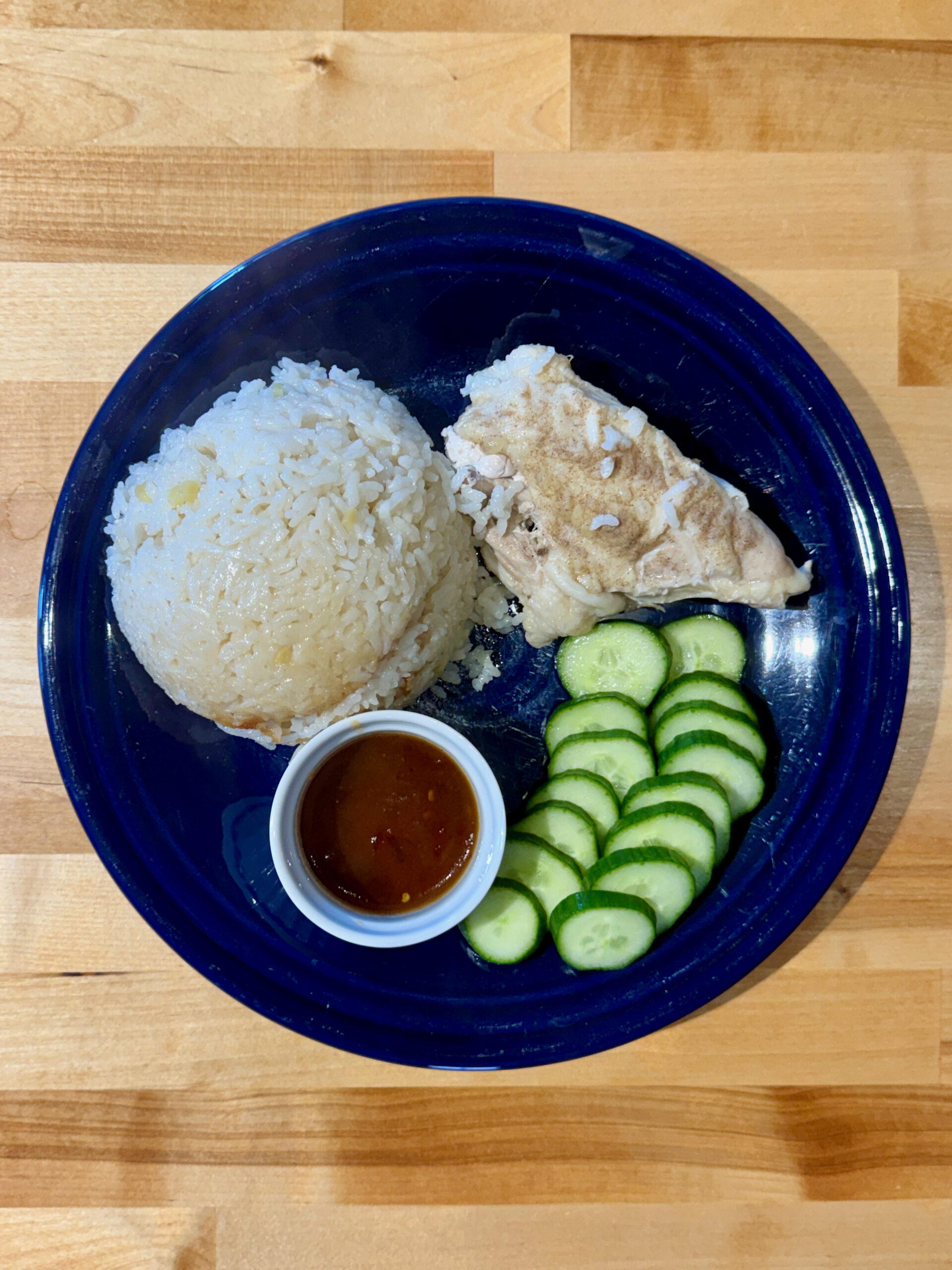 Hiananese chicken breast with cooked rice, sliced cucumber and a small white ramekin of sweet and spicy sauce on a blue plate
