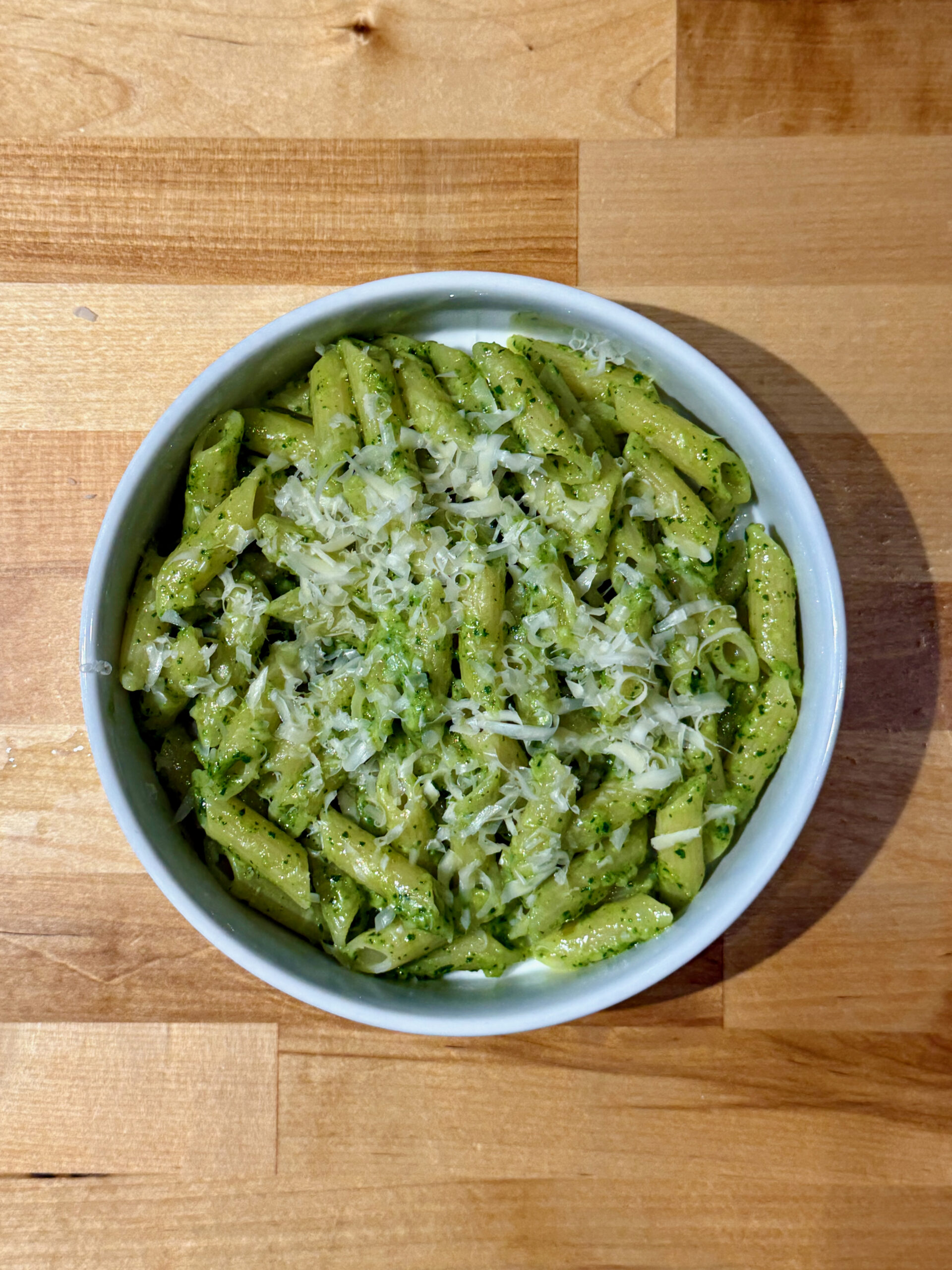 White bowl with cooked penne pasta sauced with parsley pesto and topped with grated parmesan cheese