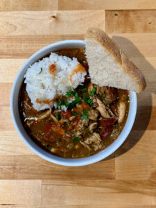 White bowl filled with chicken and sausage gumbo with chopped parsley, half a cup of cooked white rice, a quarter slice of French bread and Louisana hot sauce on top.