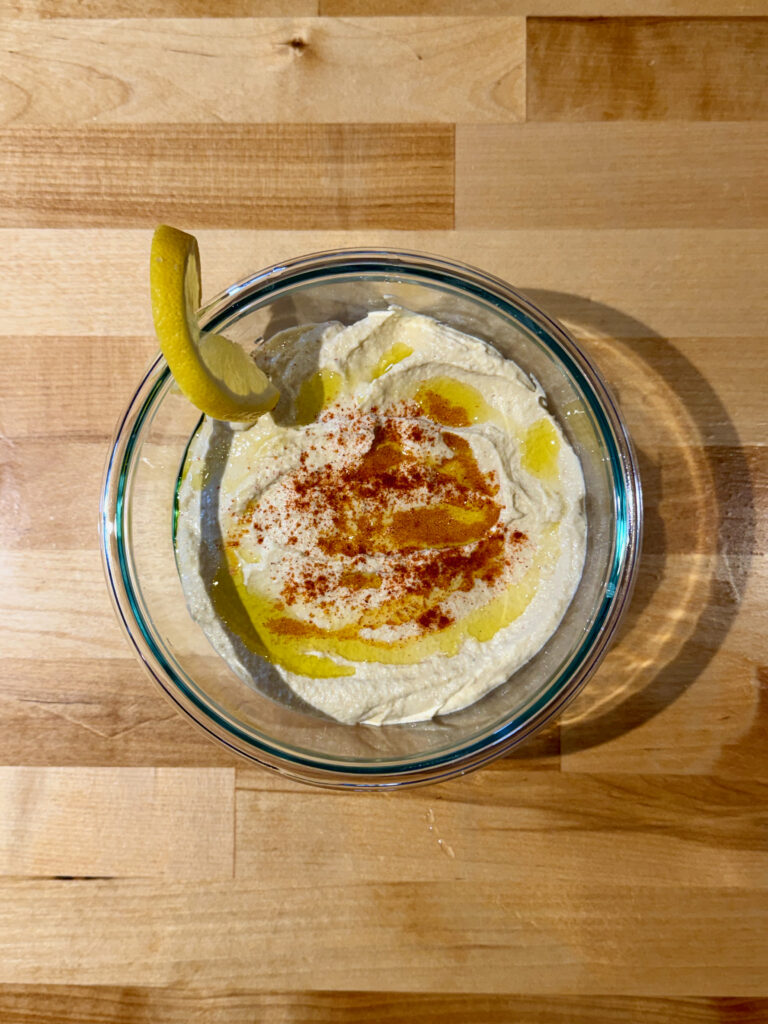 Fresh hummus in a glass bowl with a drizzle of olive oil, a sprinkle of smoked paprika, and a lemon wheel
