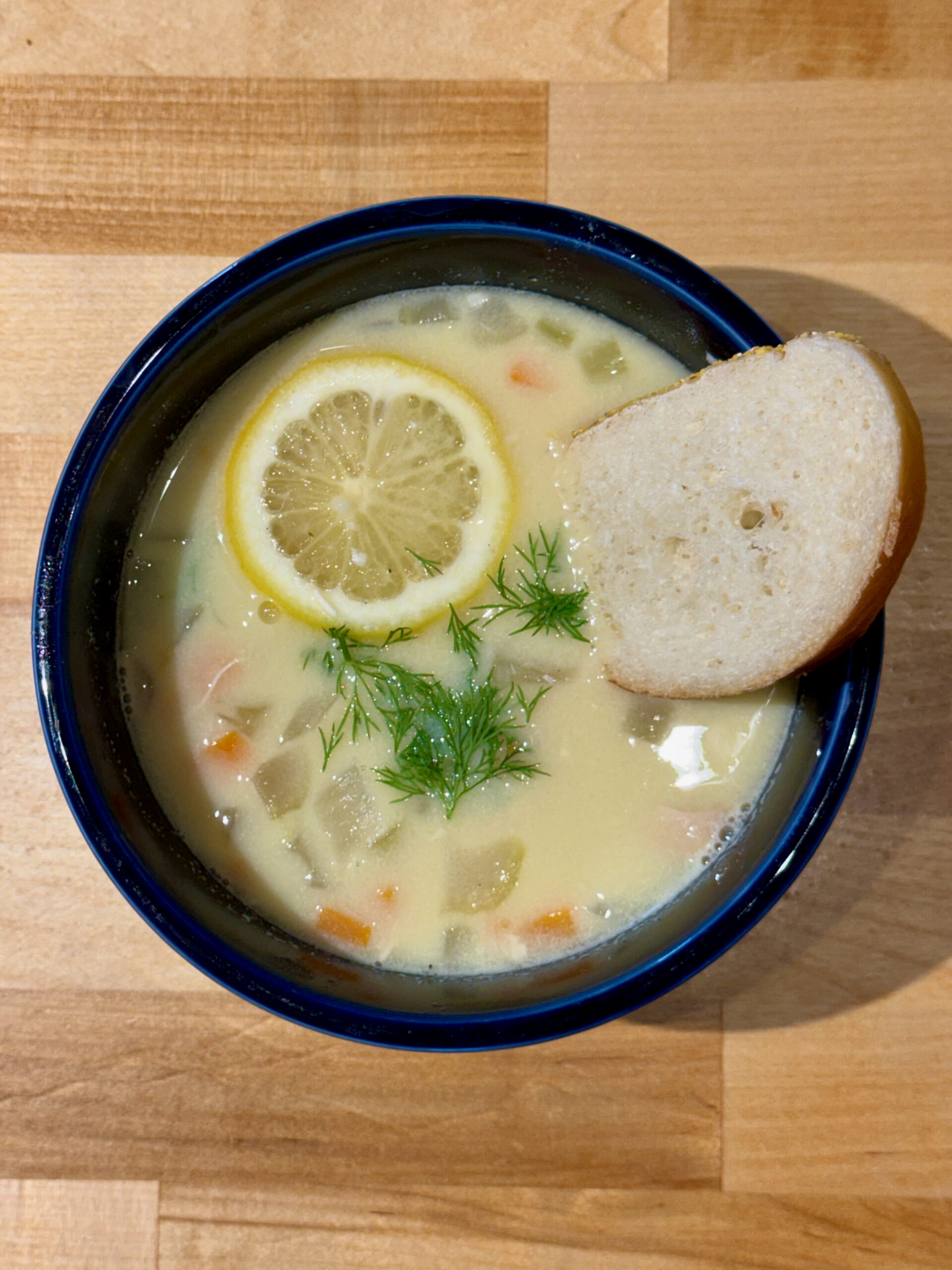 Bowl of Greek Lemon Chicken soup in a blue bowl with a pinch of fresh dill and a slice of French bread.