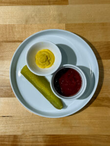 Yellow mustard and red ketchup each in white serving dishes with a pickle spear on a small white plate