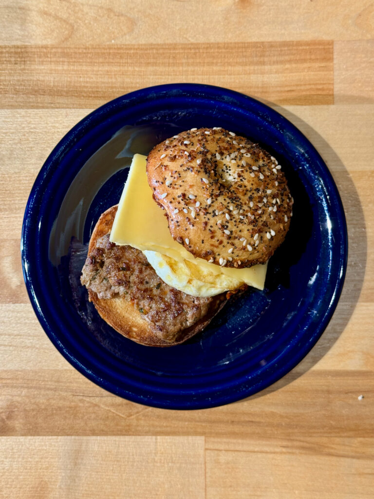 Toasted No Salt Everything Bagel with homemade breakfast sausage, egg and a slice of gouda cheese on a blue plate