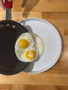 Two fried eggs being plated onto a white plate from a non-stick skillet